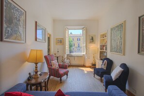 A cozy living room with a view of a building through an open window, a lamp, a chair, and a sofa.