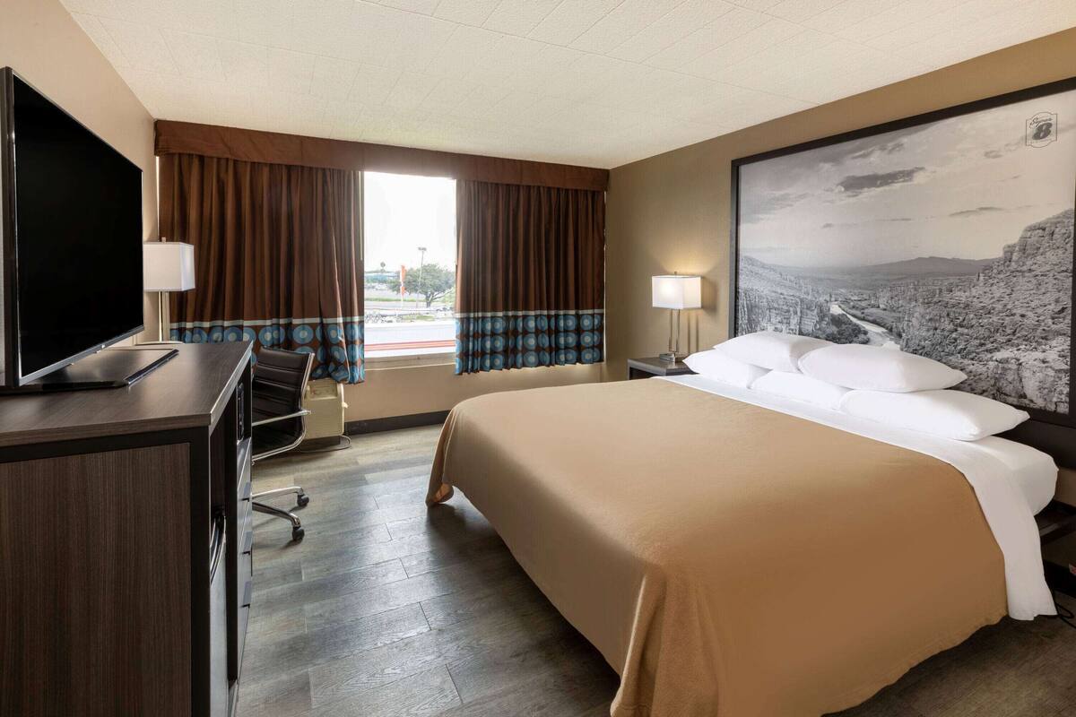 A hotel room with a bed, a desk with a TV, a chair, a window with curtains, and a painting on the wall.