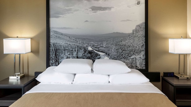A hotel room with a large framed black and white photograph of a mountainous landscape on the wall, a neatly made bed with white linens, two bedside tables with lamps, and a digital clock displaying the time. - Super 8 by Wyndham Laredo (Laredo)