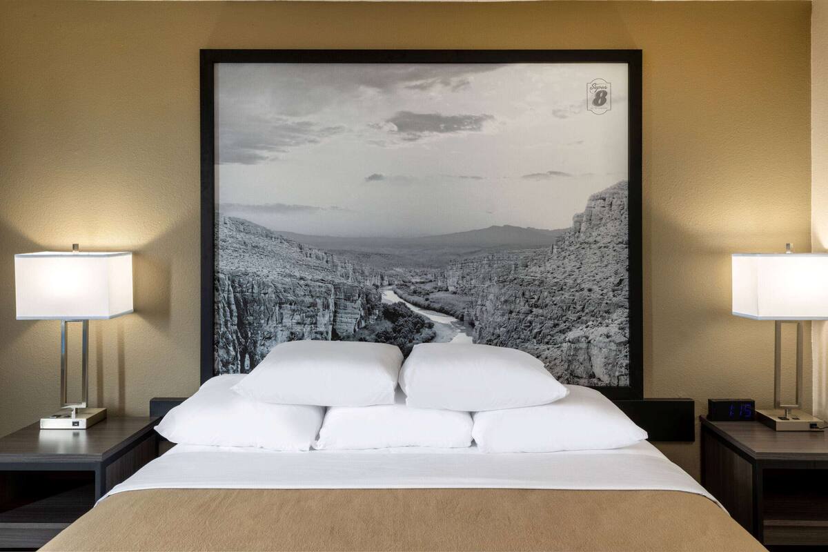 A hotel room with a large framed black and white photograph of a mountainous landscape on the wall, a neatly made bed with white linens, two bedside tables with lamps, and a digital clock displaying the time.