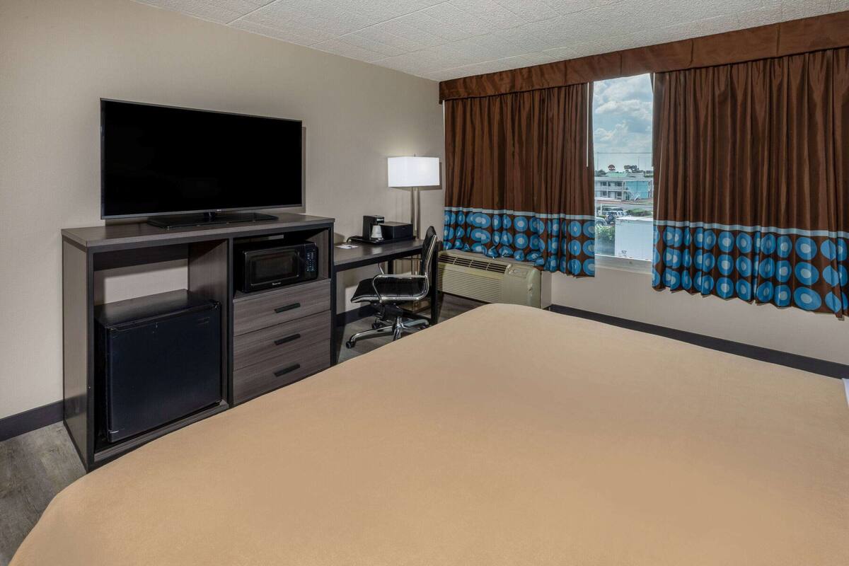 A hotel room with a large bed, a desk with a flat-screen TV, a mini-fridge, a lamp, and a view of the city through the window.