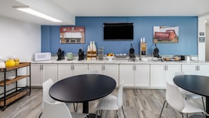 A modern kitchen with white cabinets, a blue accent wall, and a flat-screen TV. There are two black dining tables with white chairs. - Super 8 by Wyndham Laredo (Laredo)