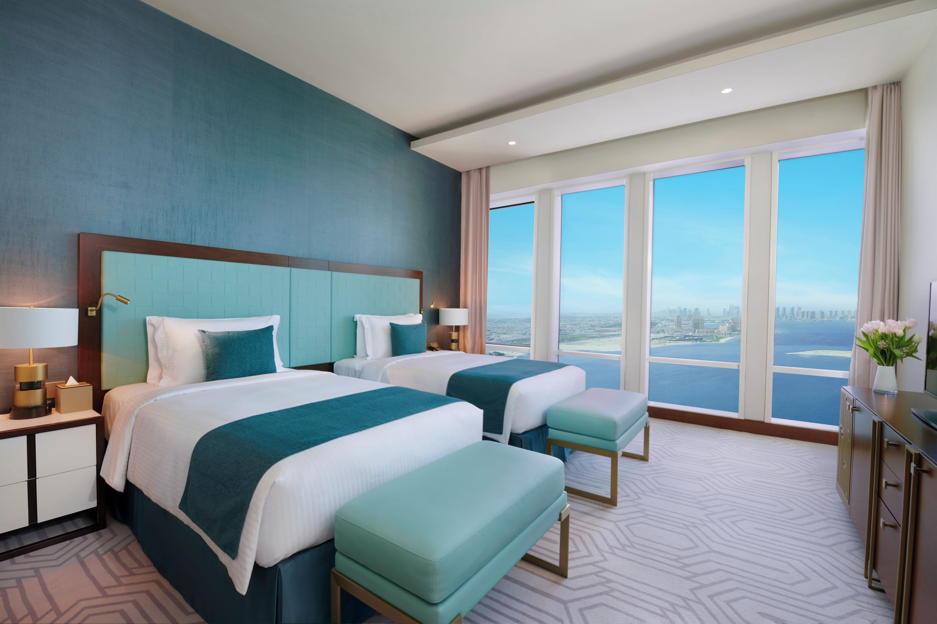 A hotel room with two beds, a large window with a city view, a desk with a lamp, and a nightstand with a lamp and a box.