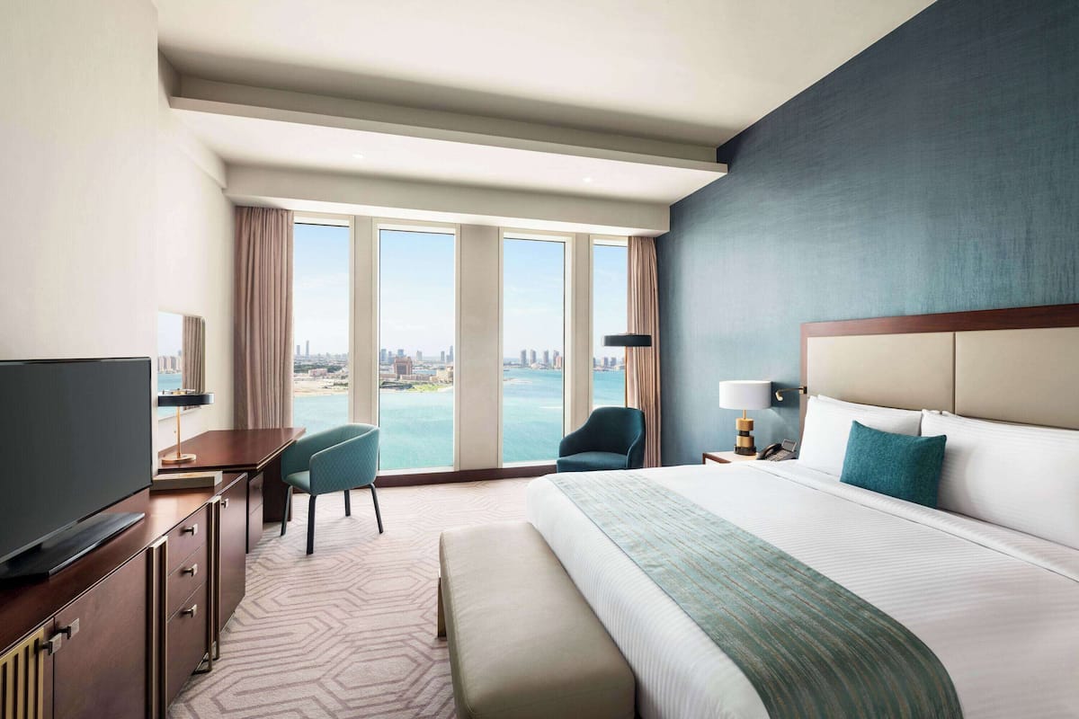 Ein Hotelzimmer mit einem großen Bett, einem Schreibtisch mit Fernseher, einem Sessel und Blick auf die Stadt durch große Fenster.