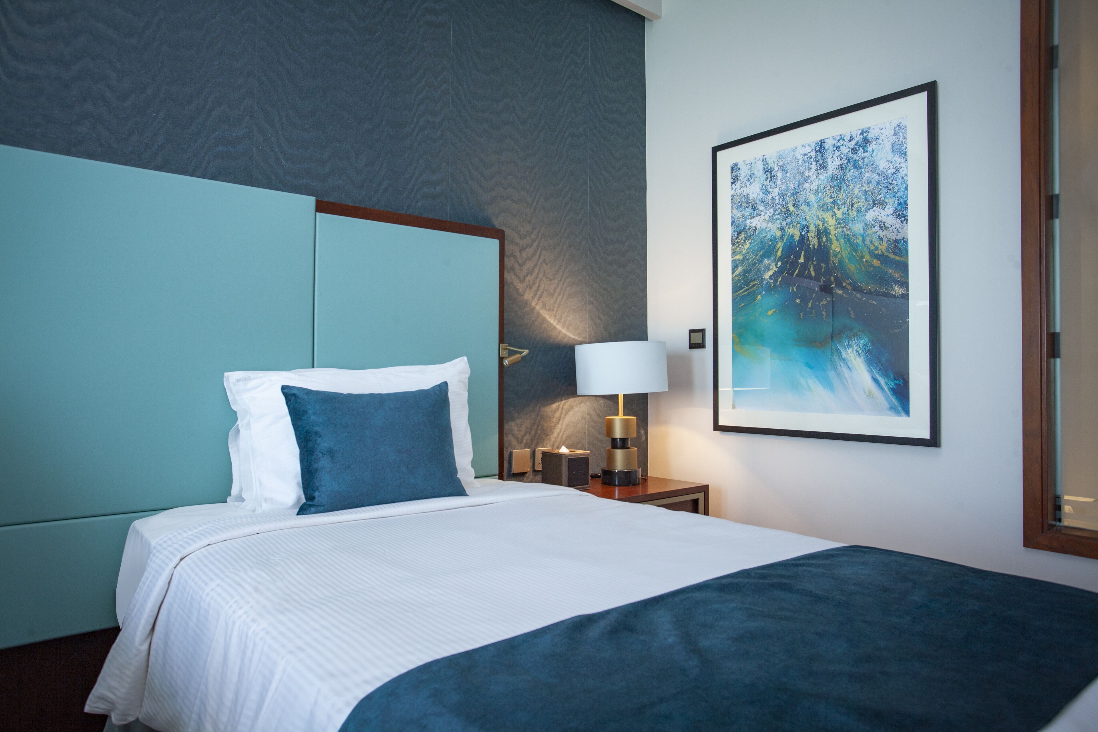 A neatly made bed with white and blue bedding, a bedside table with a lamp, and a framed abstract painting on the wall.