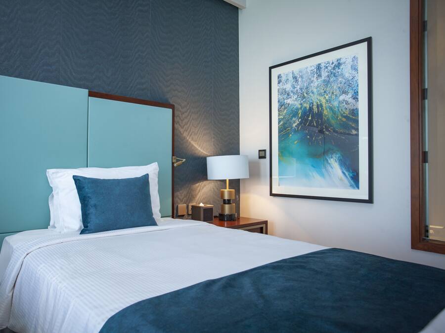 Cama bem arrumada com roupa de cama branca e azul, uma mesa de cabeceira com abajur e um quadro abstrato na parede.