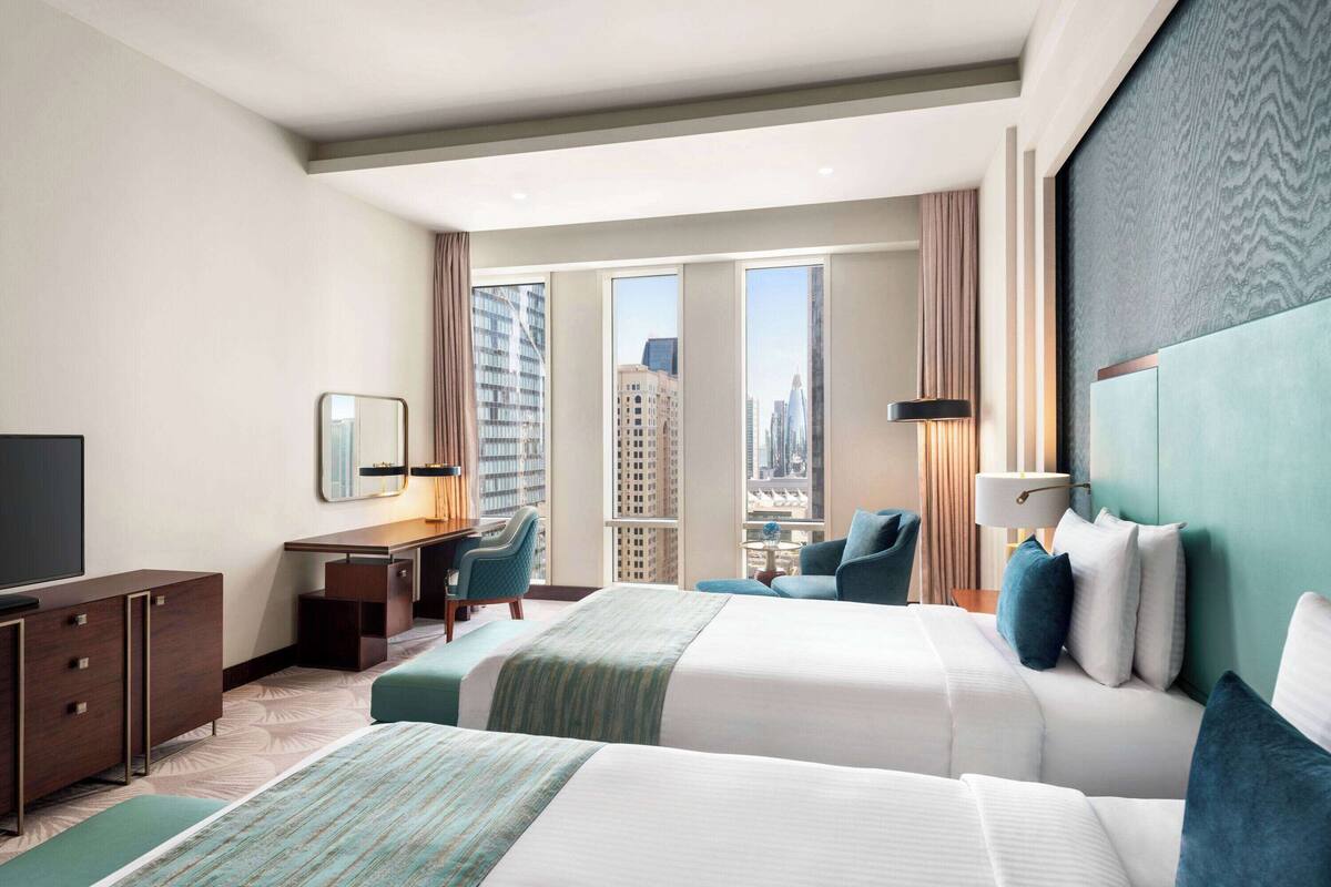 Ein Hotelzimmer mit zwei Betten, einem Schreibtisch, einem Stuhl, einem Fernseher und einem großen Fenster mit Stadtblick.