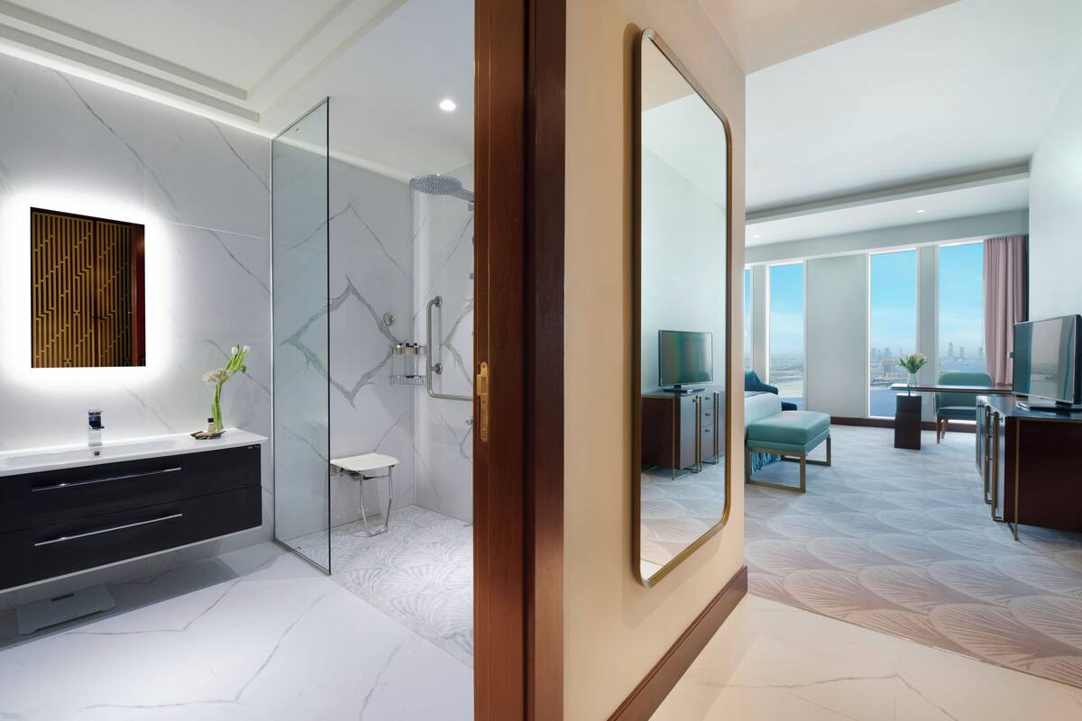Ein modernes Hotelzimmer mit großem Spiegel, begehbarem Kleiderschrank, Badezimmer mit Glasdusche und Blick auf die Stadt durch große Fenster.