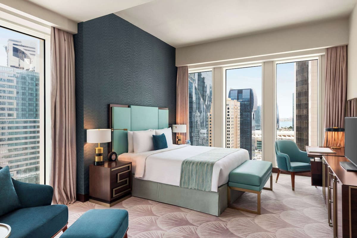 Ein modernes Hotelzimmer mit einem großen Bett, einem Schreibtisch und einem Stuhl. Vor den Fenstern hängen Vorhänge, und das Zimmer bietet Blick auf die Stadt.