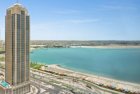 Un immeuble imposant à l’architecture moderne, situé à côté d’une plage aux eaux d’un bleu limpide et dotée d’une promenade bien entretenue.