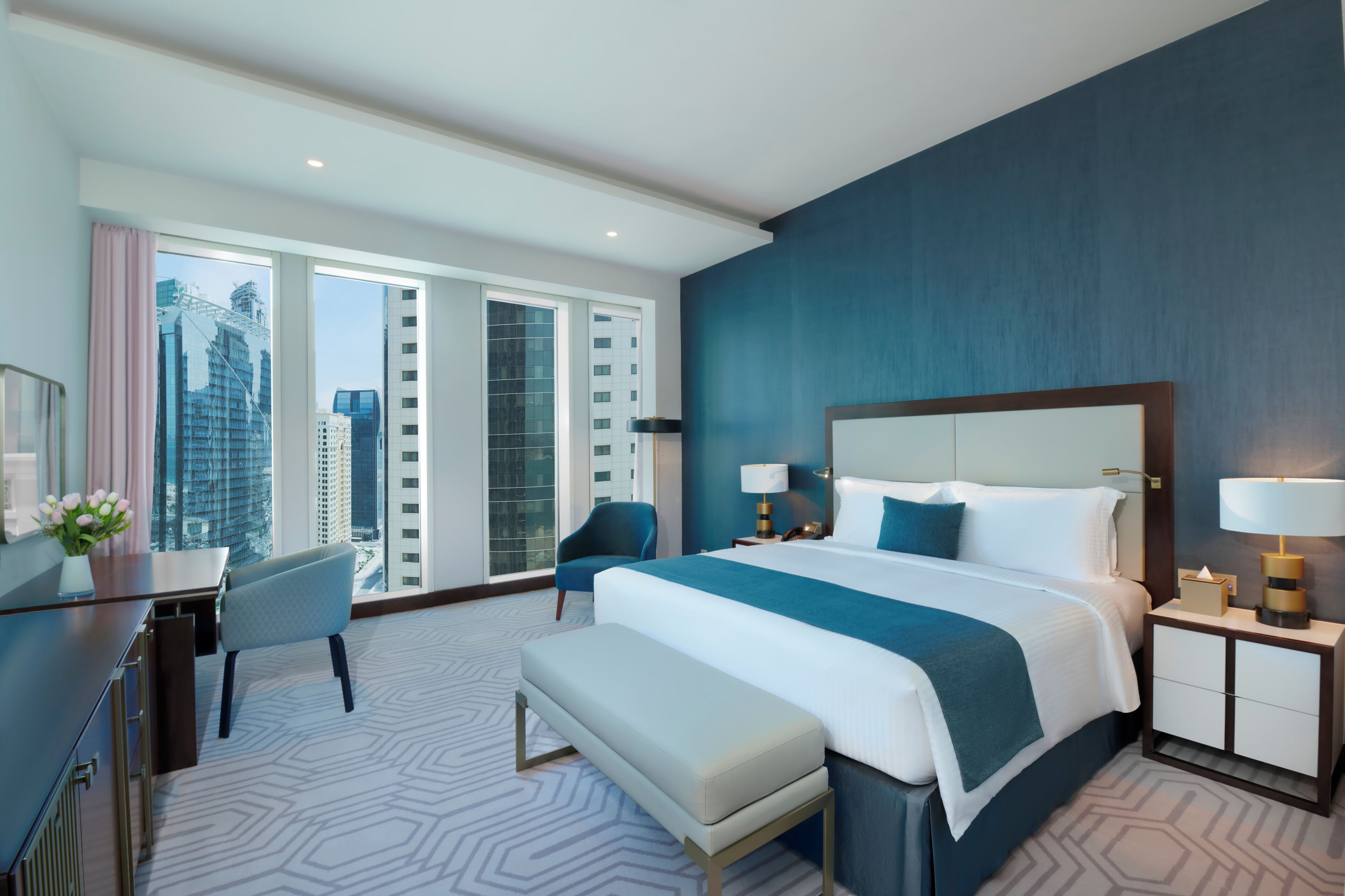 Ein modernes Hotelzimmer mit einem großen Bett, einem Schreibtisch, einem Stuhl und Blick auf die Stadt durch große Fenster.