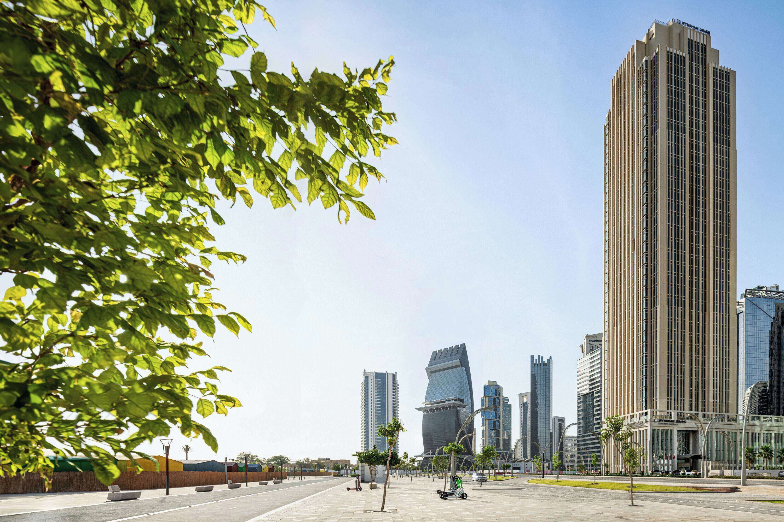 A modern urban plaza with high-rise buildings, a clear sky, and a tree-lined walkway.