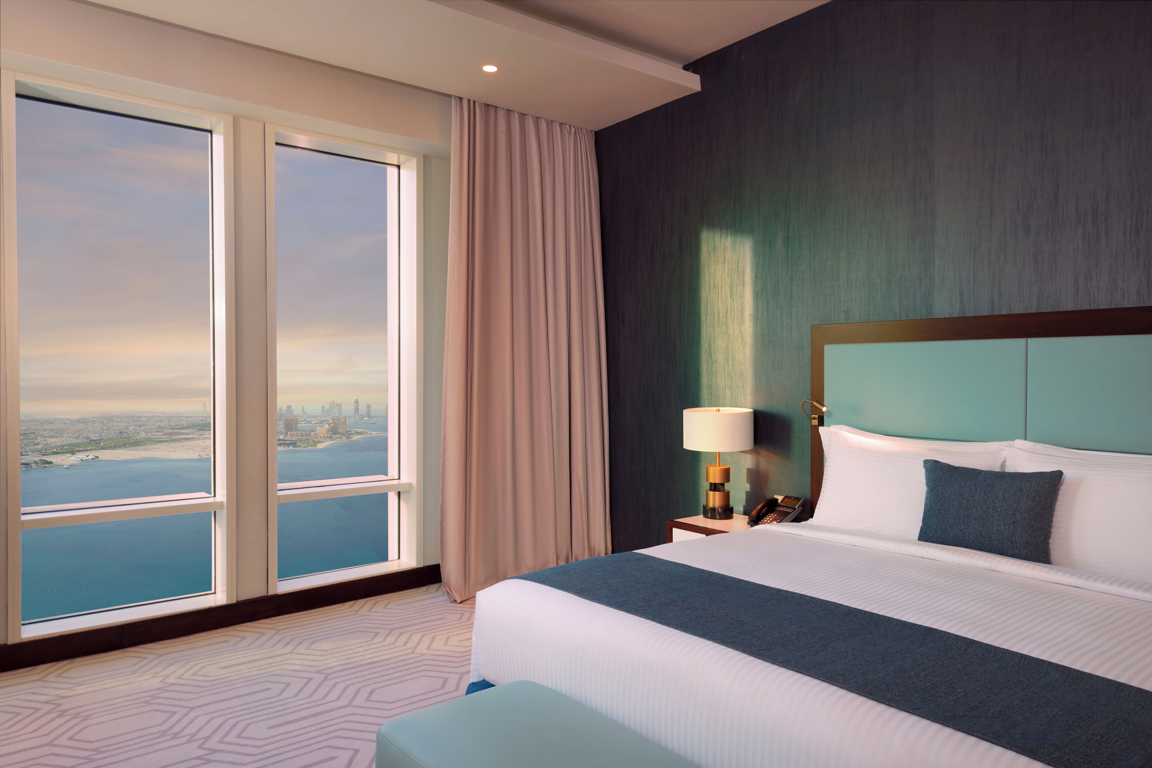Habitación de hotel con una cama grande, una mesita de noche, una lámpara y vistas de un paisaje urbano a través de ventanales de piso a techo.