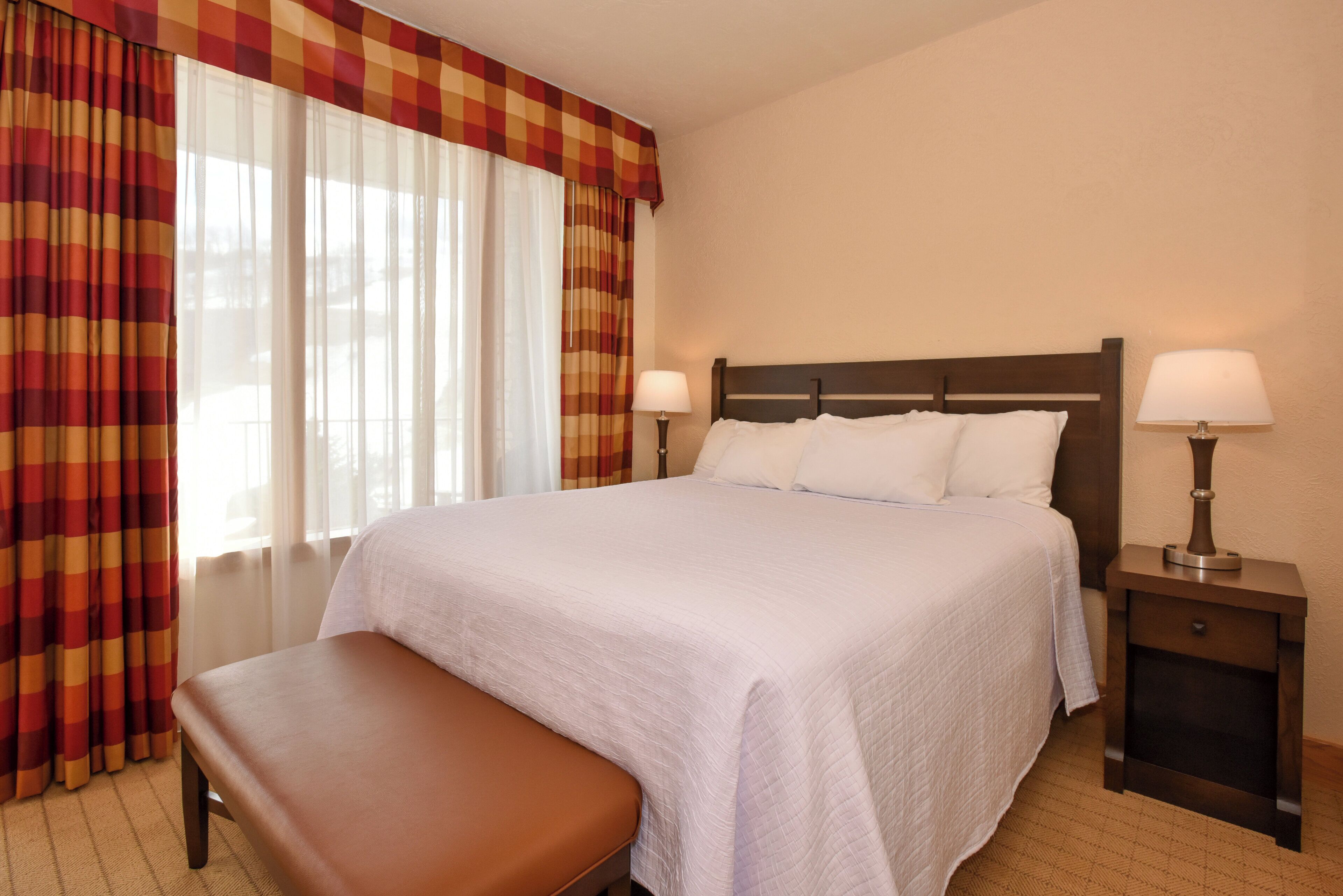 A hotel room with a large bed, two bedside lamps, a bench, and a window with curtains.