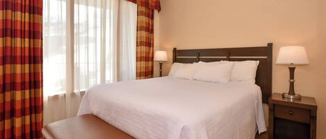 A hotel room with a large bed, two bedside lamps, a bench, and a window with curtains.