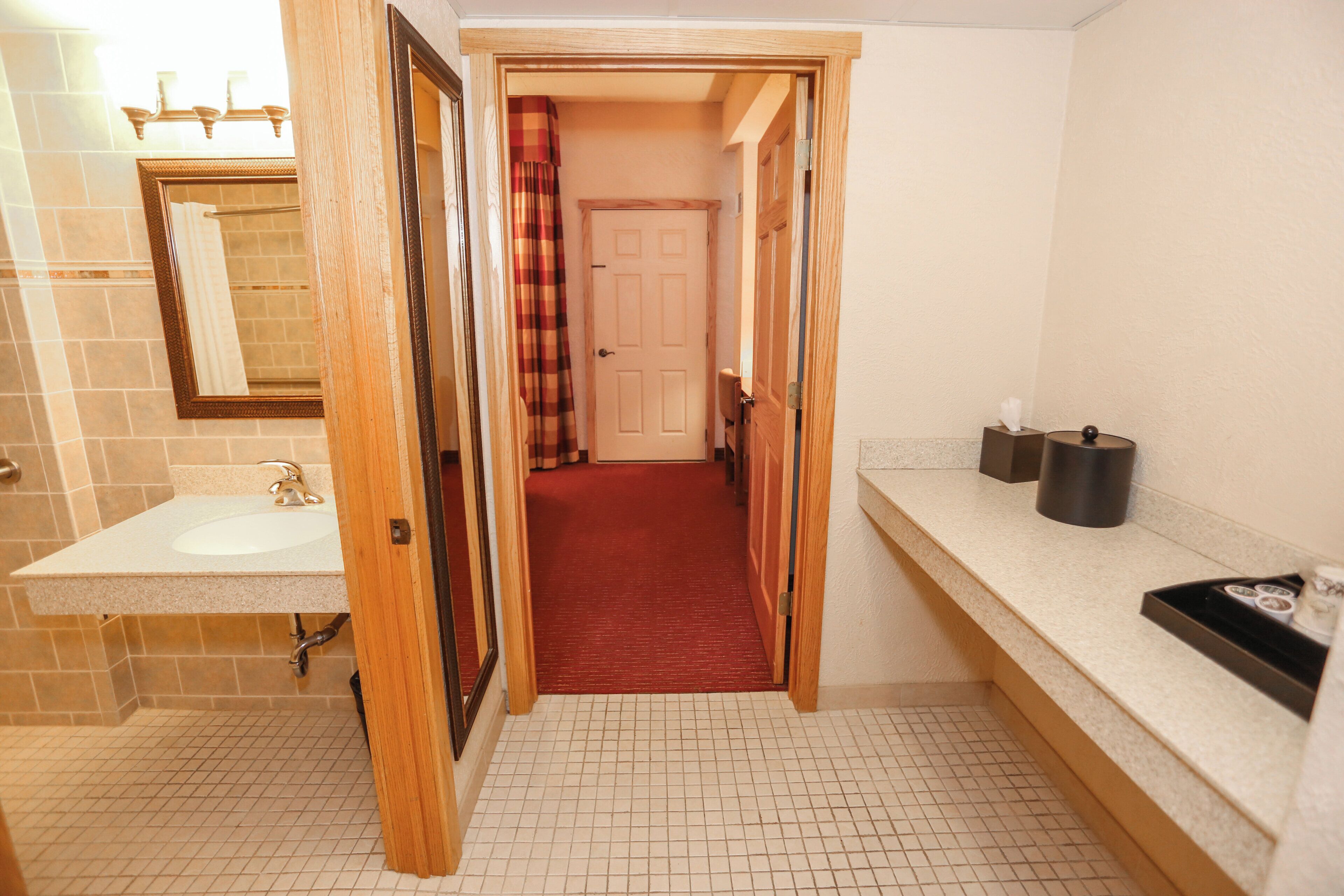 A bathroom with a sink, mirror, and a red carpeted hallway leading to a door.