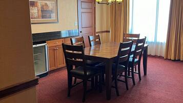 A dining area with a wooden table and chairs, a kitchen area with a refrigerator and cabinets, and a large window with curtains.