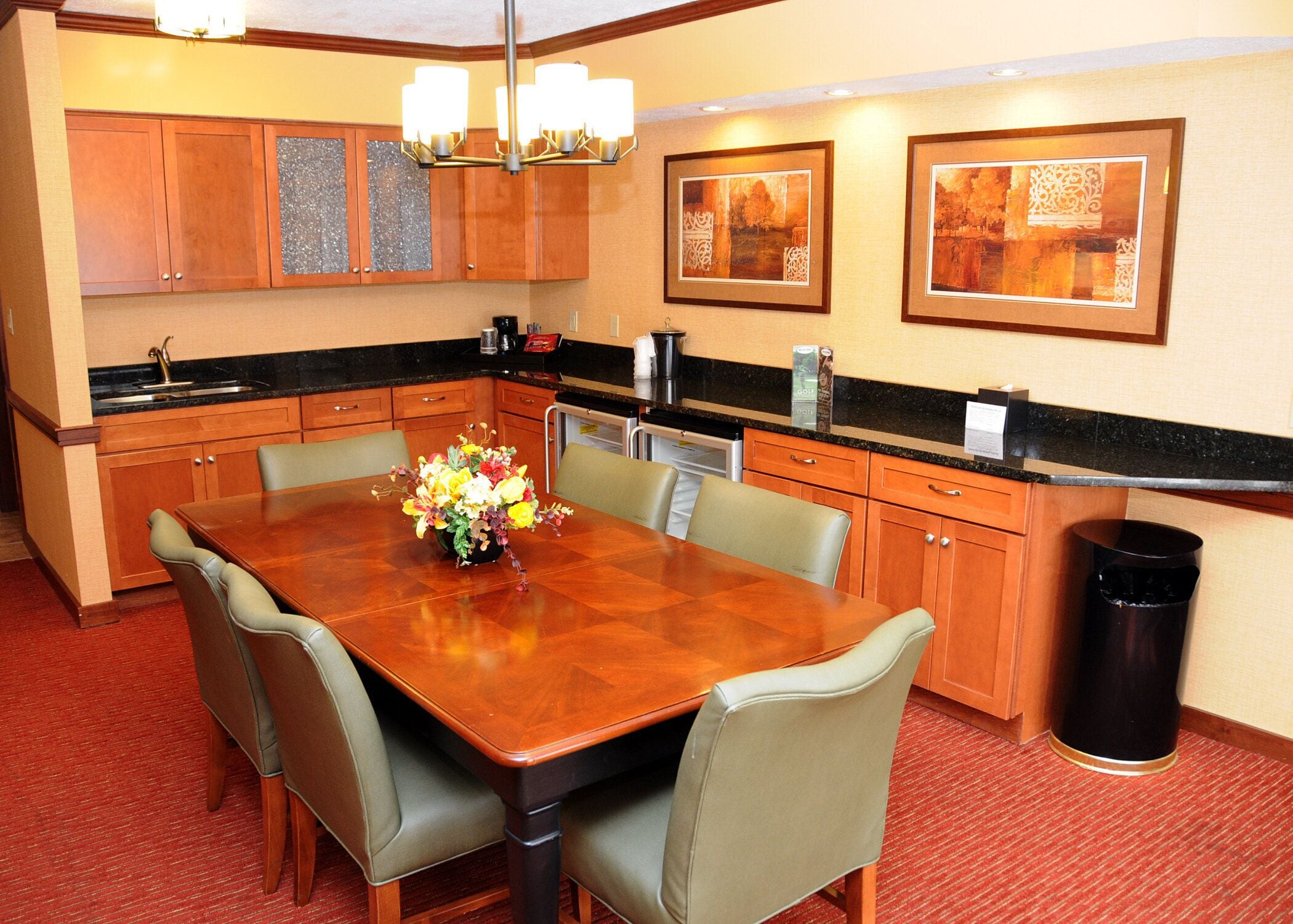 A hotel dining area with a wooden table, beige chairs, a vase of flowers, and framed artwork on the wall.
