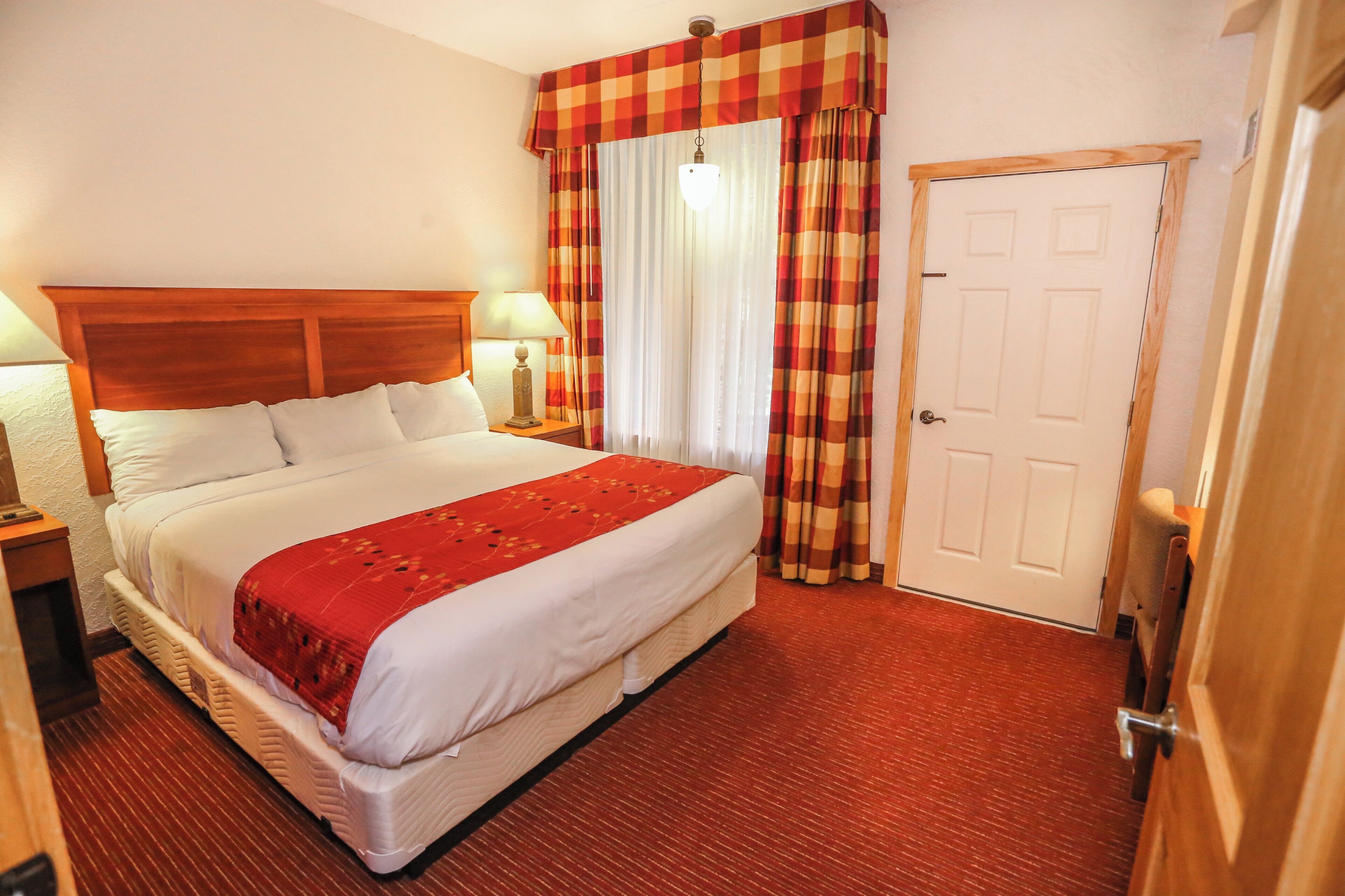 A hotel room with a large bed, a wooden headboard, two bedside tables with lamps, a red carpeted floor, and a checkered curtain over the window.