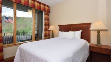 A neatly made bed with white bedding, two bedside lamps, and a window with a view of a green landscape and an American flag.