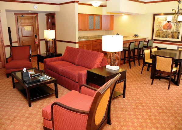 Habitación de hotel con sala de estar que incluye un sofá, sillones, una mesa de centro y una mesa de comedor con sillas. Cuenta con una zona de cocina con armarios de madera y una encimera.