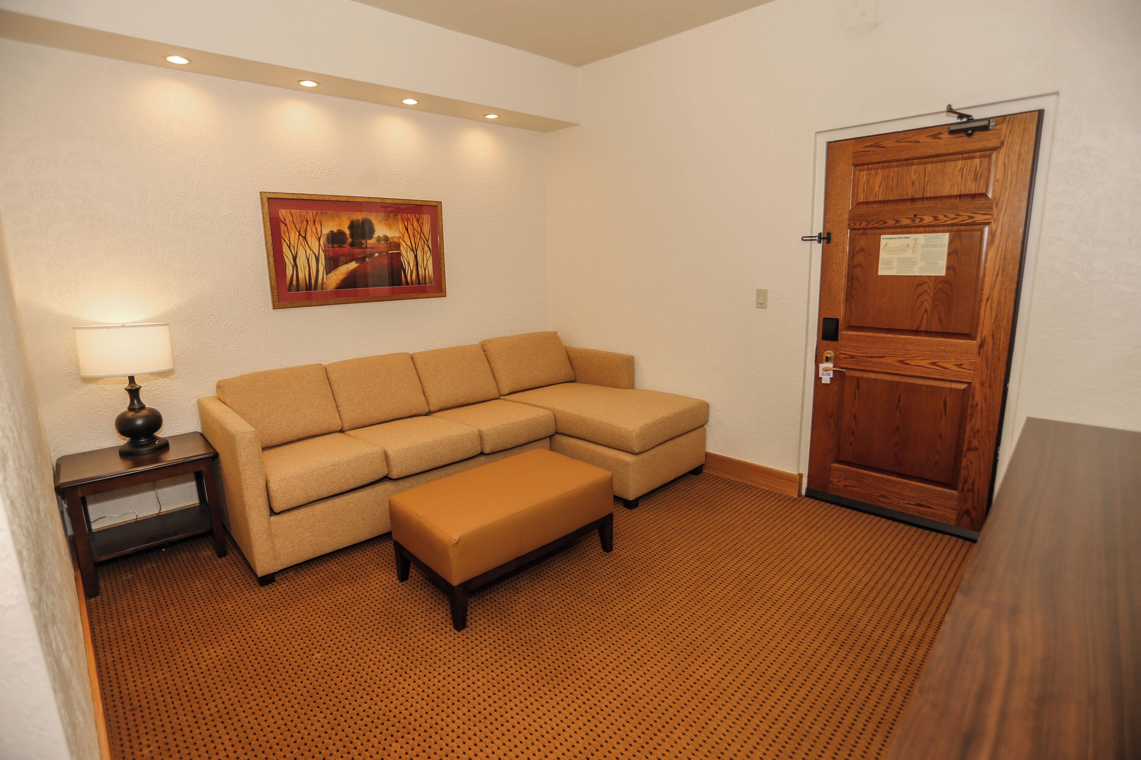 A room with a beige sectional sofa, a wooden door, a framed picture on the wall, and a lamp on a side table.