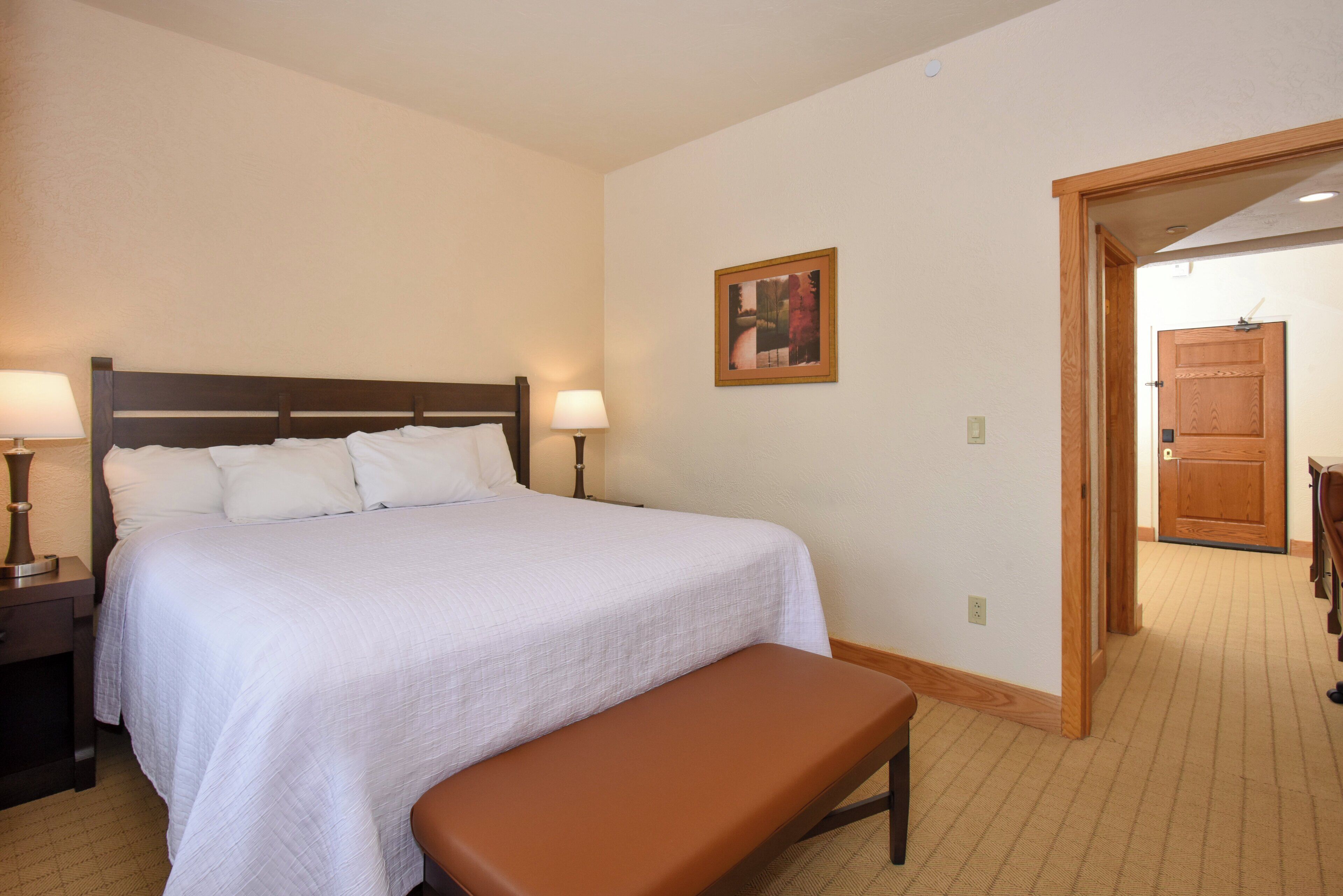A hotel room with a large bed, two bedside lamps, a wooden bench, a framed picture on the wall, and a wooden door.