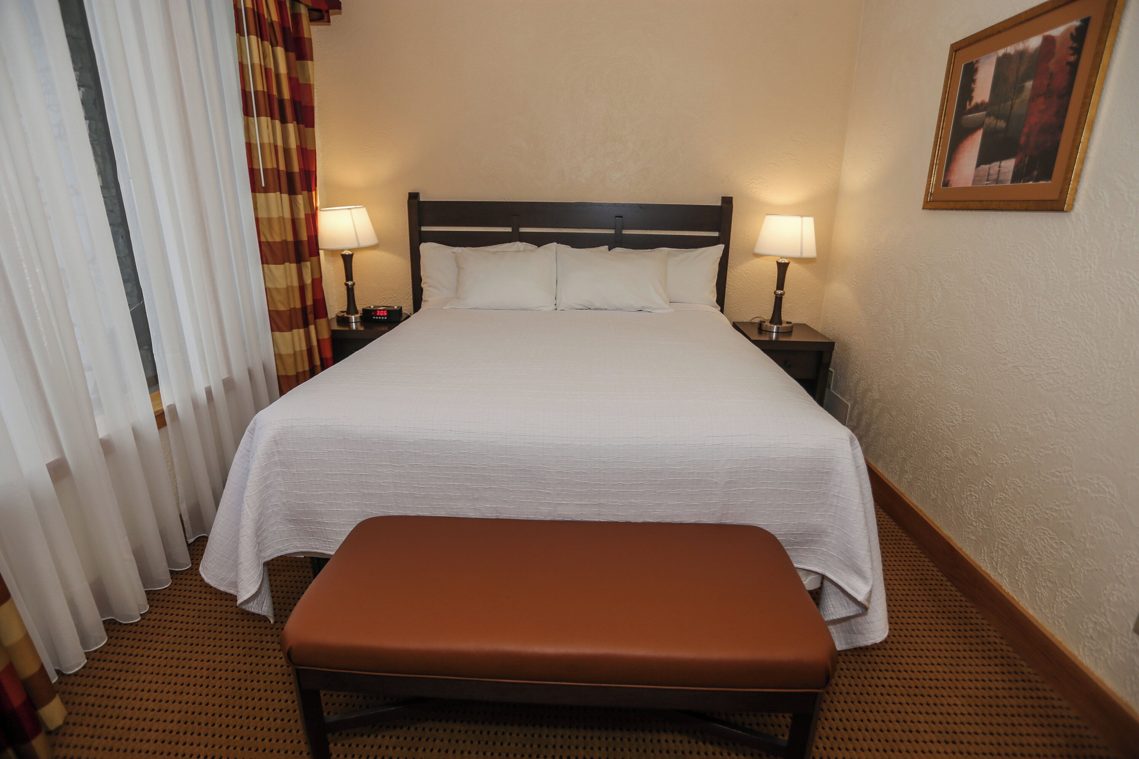 Une chambre d’hôtel avec un grand lit, deux tables de chevet avec des lampes, un banc et une fenêtre avec des rideaux.