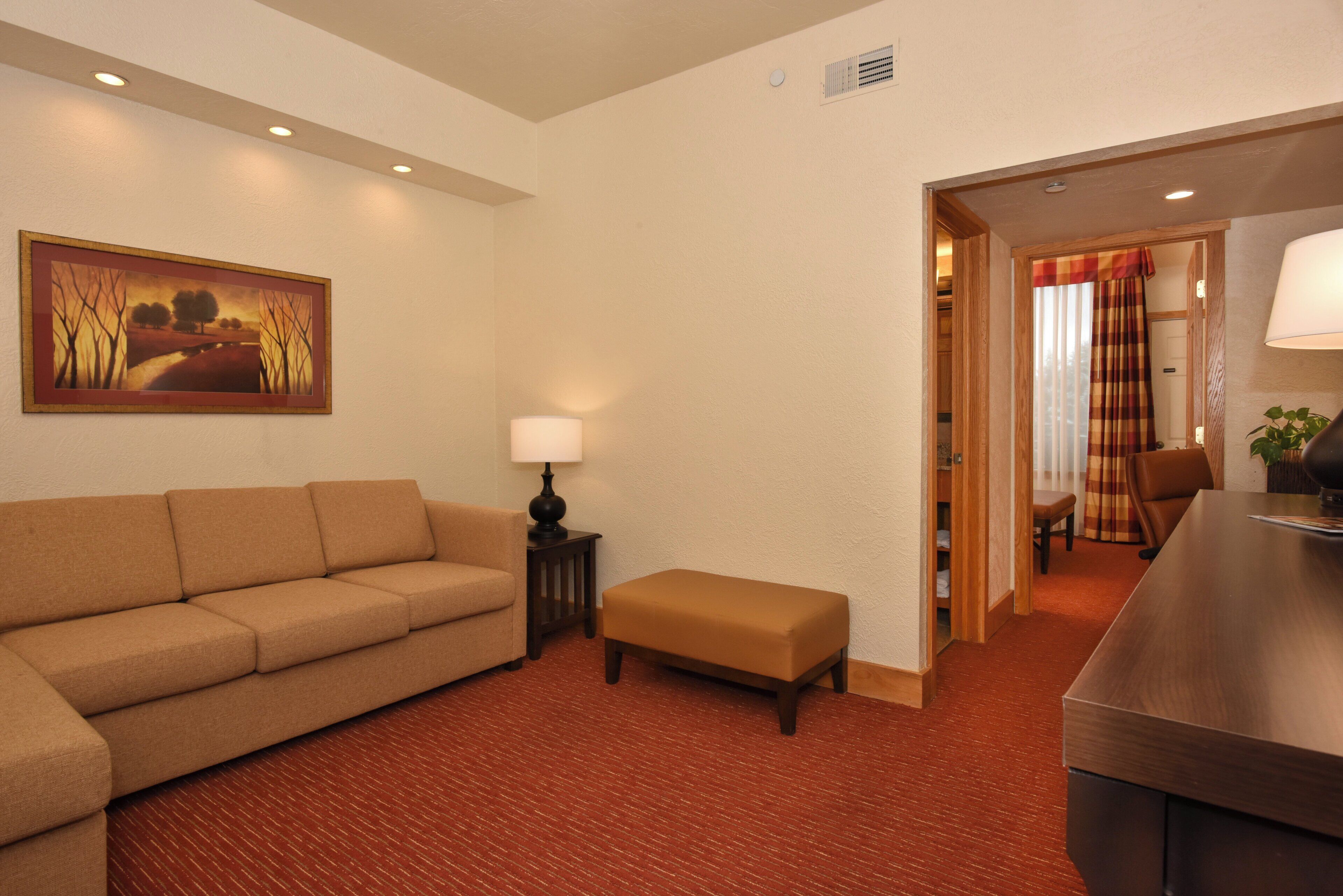 A hotel room with a sofa, an ottoman, a lamp, a painting, and a wooden desk.