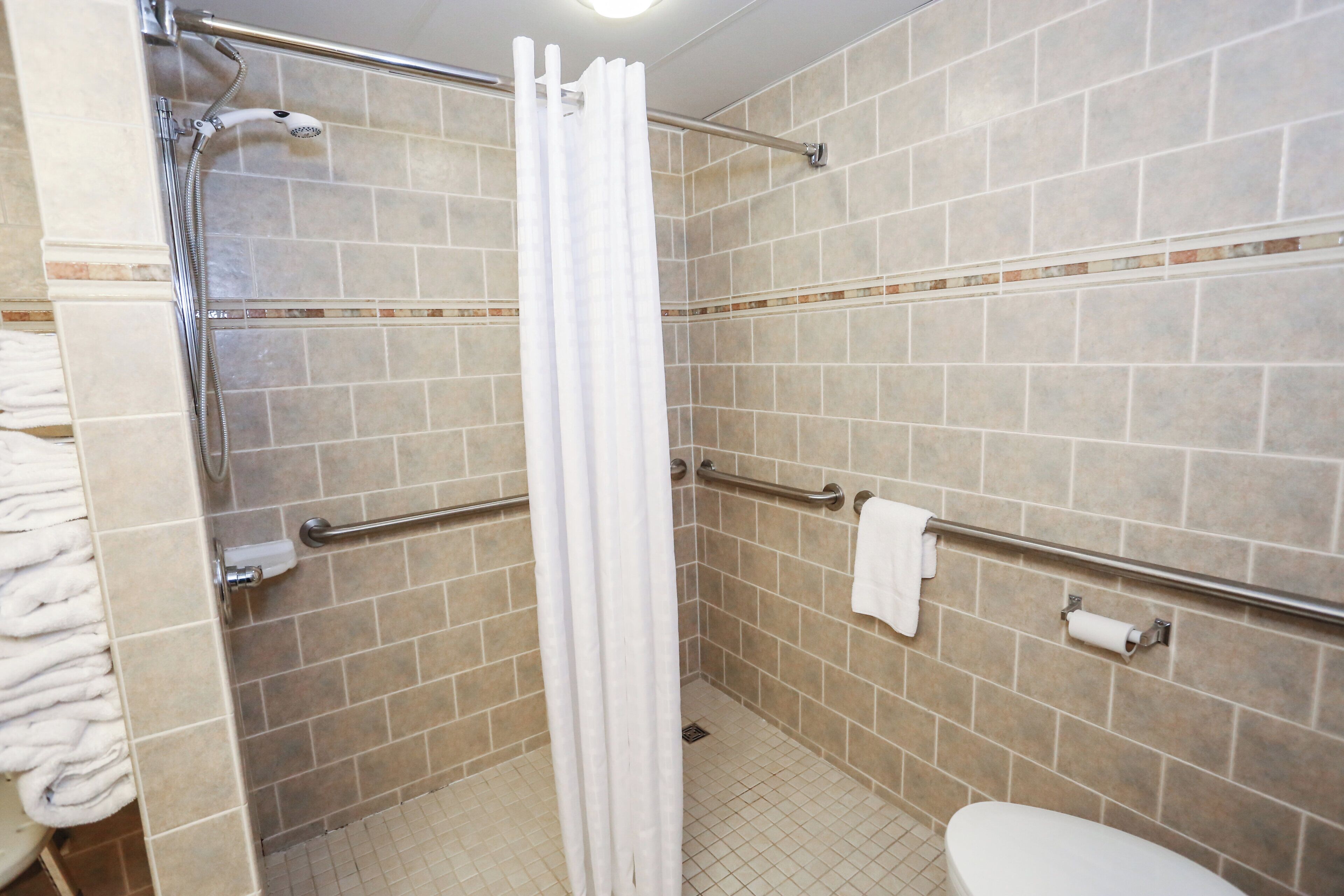 Un baño con azulejos color crema, un inodoro blanco, una ducha con cortina blanca y barras de apoyo.