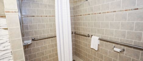 Et badeværelse med beige fliser, et hvidt toilet, en bruser med hvidt gardin og gelændere.