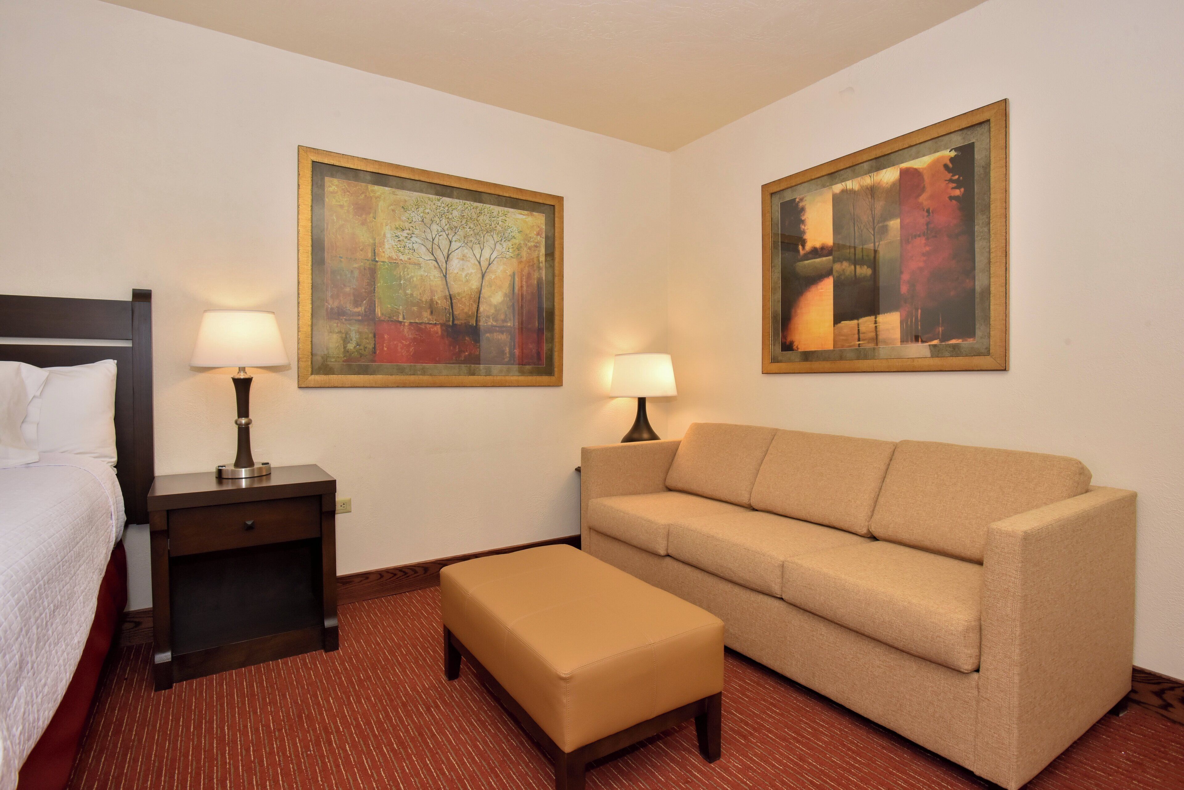 A hotel room with a bed, a sofa, an ottoman, two nightstands with lamps, and two framed paintings on the walls.