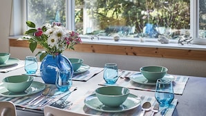 Ein Esstisch ist gedeckt mit grünen Schüsseln, Tellern und Besteck, dazu eine blaue Vase mit Blumen und eine hängende Leuchte.