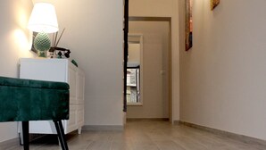 Een gang met een groene bekleden bank, een wit kastje en een lamp. Er hangen kunstwerken aan de muur en bevindt zich een deur aan het einde van de gang.