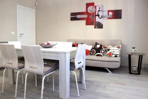 A dining area with a white table and chairs, a grey sofa with patterned cushions, and abstract wall art.