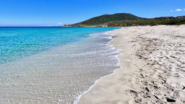 En strand med kristallklart turkost vatten, en sandstrand och en kulle i bakgrunden.
