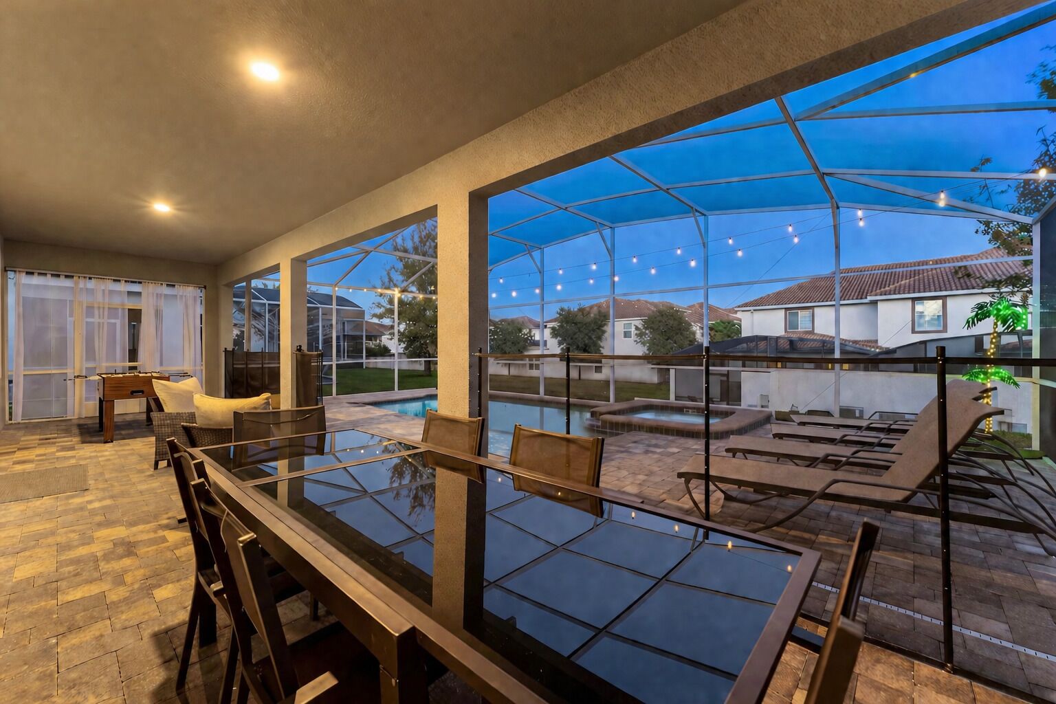 An outdoor dining area with a glass table and chairs, surrounded by a covered patio with string lights.