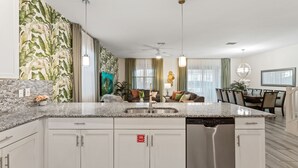Una cocina moderna con armarios blancos, encimera de granito y electrodomésticos de acero inoxidable. Cuenta con una zona de comedor con mesa y sillas, además de una sala de estar con sofá y un cuadro en la pared.