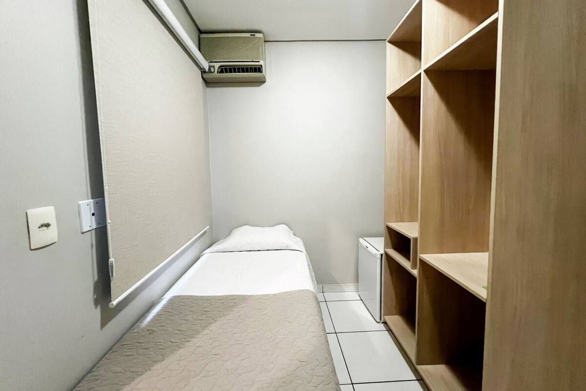A small, narrow room with a bed, a wooden wardrobe, and a wall-mounted air conditioner.