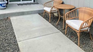 A patio area with two wicker chairs and a small table, a concrete pathway leading to a sliding glass door, and a blue gas cylinder on the ground.