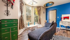 Une pièce avec un canapé à motifs, une malle verte, une petite table ronde et une fenêtre avec des rideaux.