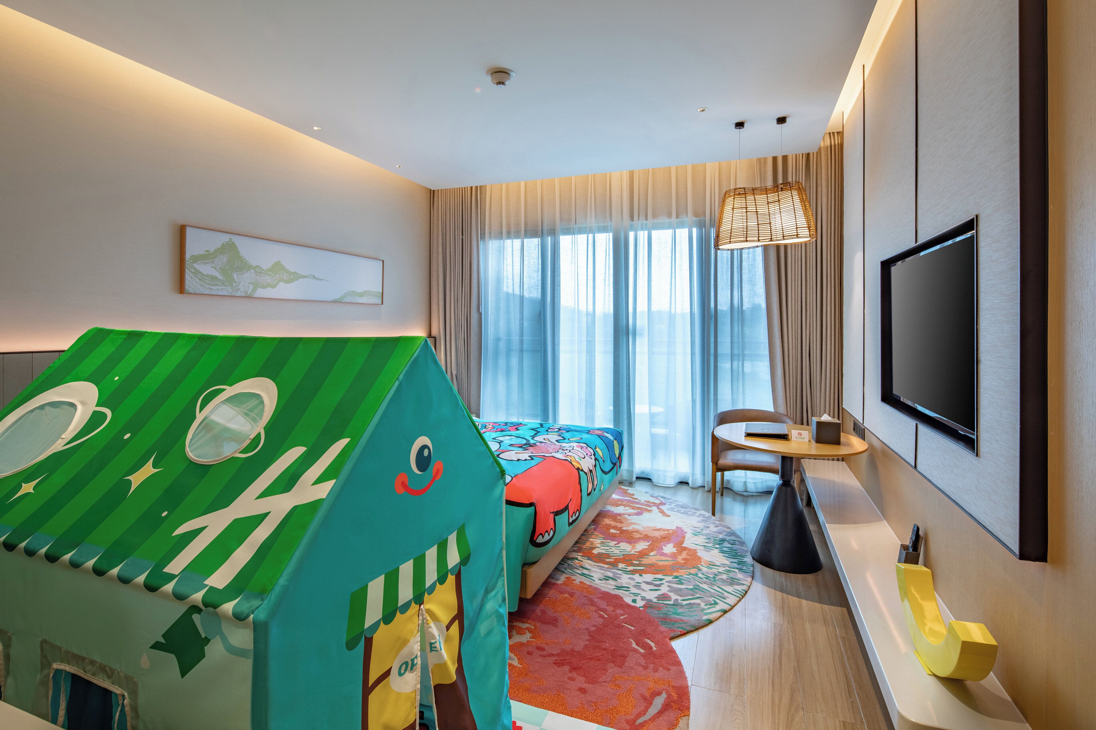 Ein modernes Hotelzimmer mit einem großen Bett, einem Fernseher, einem Spielhaus und Blick ins Freie durch das Fenster.