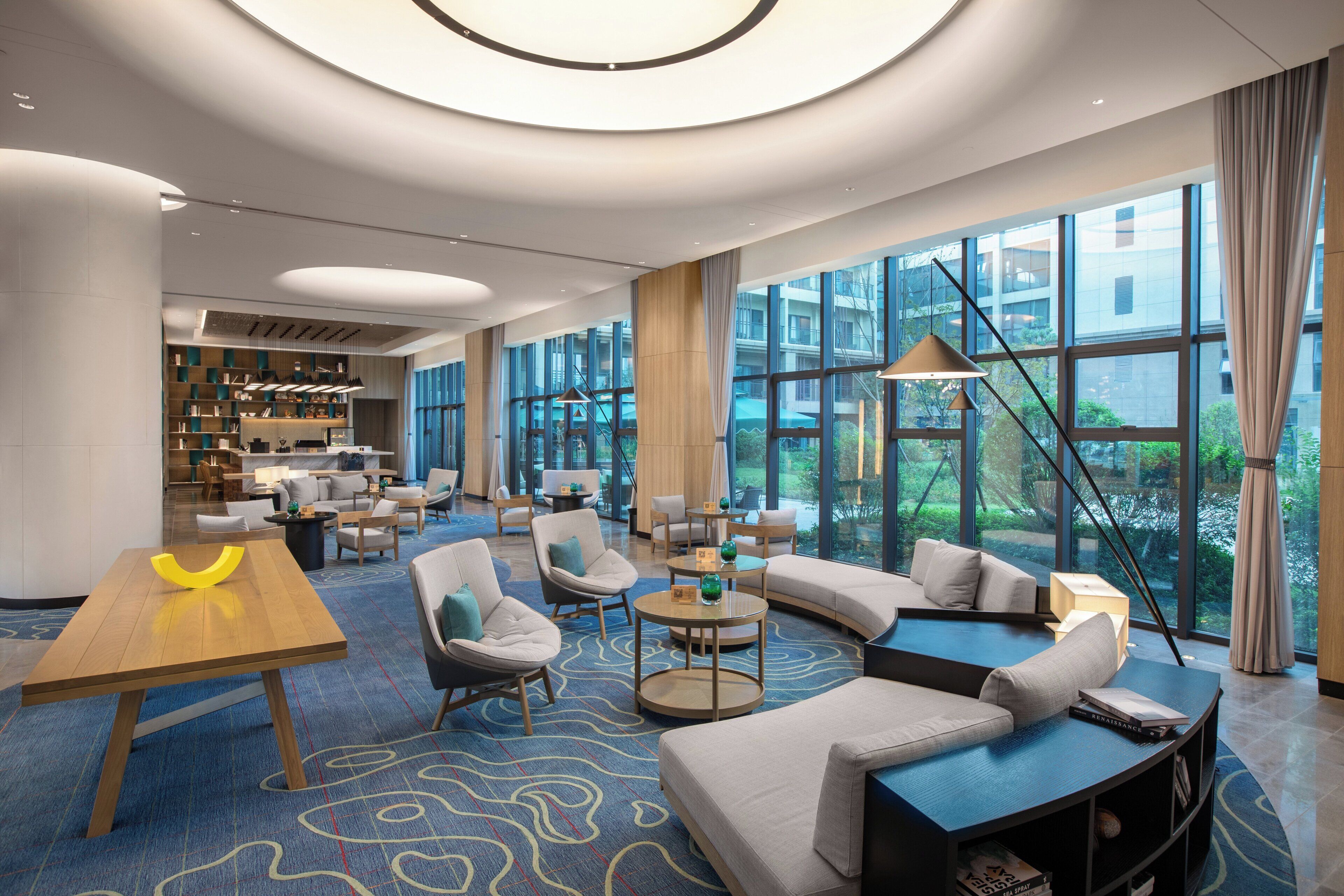 A spacious lobby with a curved sofa, round tables, and a wooden coffee table. Large windows offer a view of the outdoors.