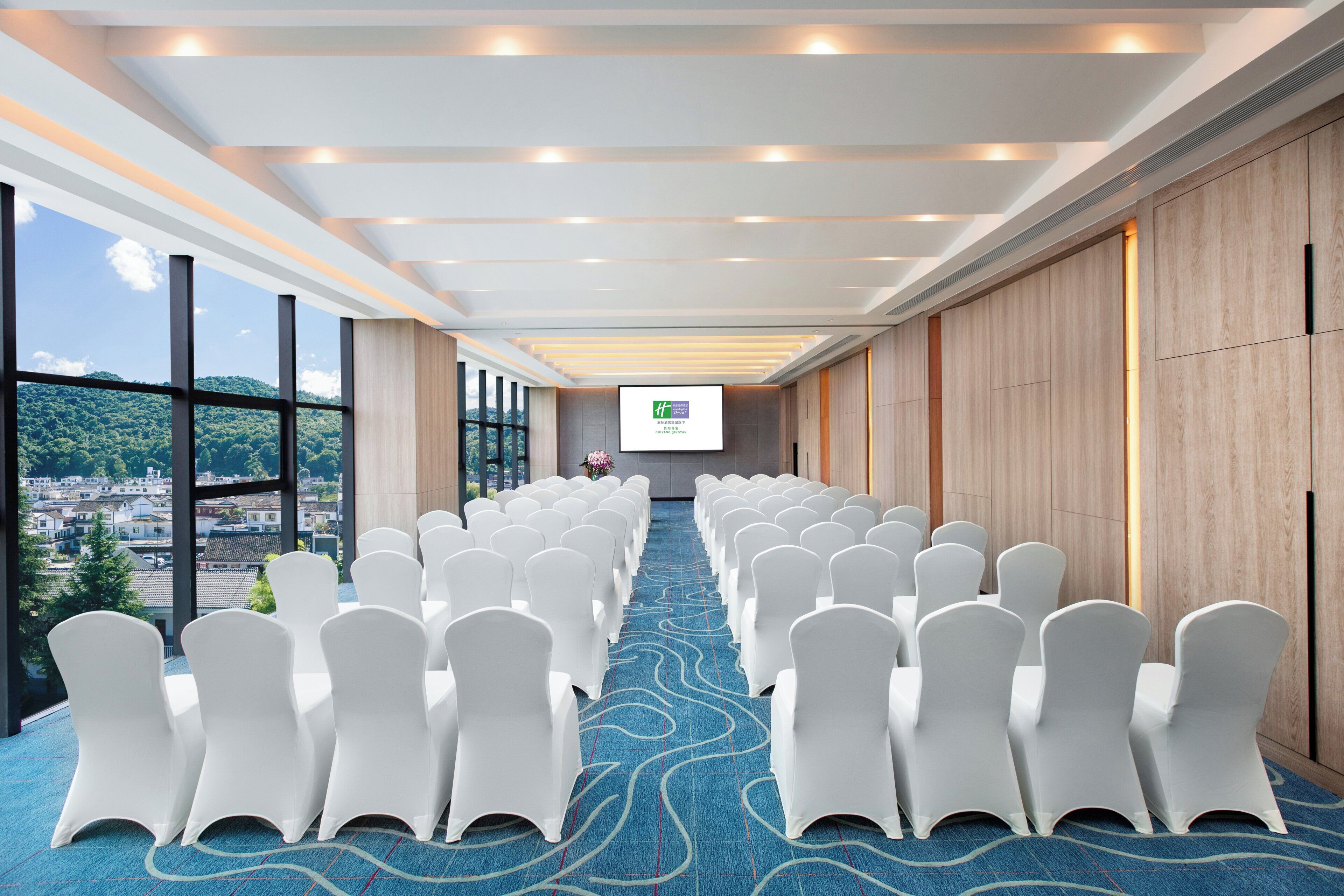 Una sala conferenze con file di sedie bianche, un tappeto blu e un grande schermo di fronte.