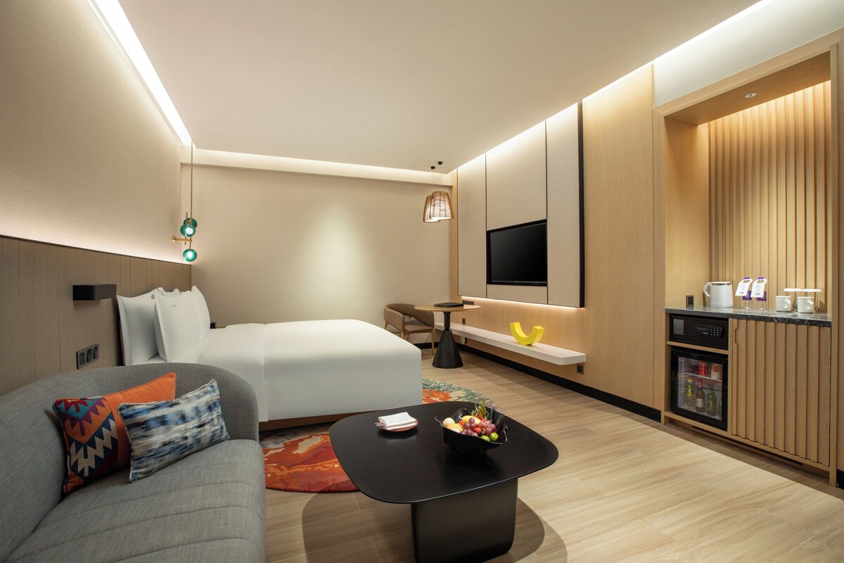 A modern hotel room with a bed, sofa, coffee table with fruit, a TV, and a minibar.