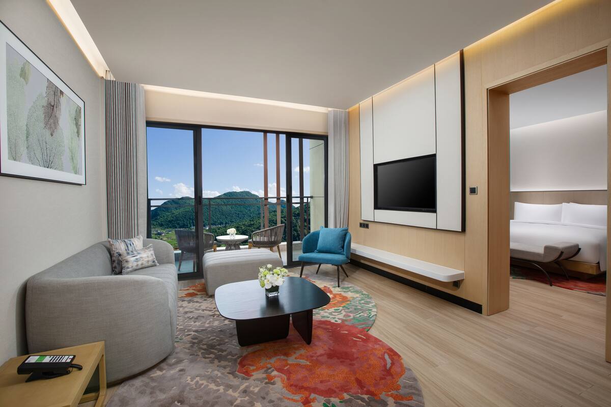 A modern hotel room with a large window, a flat-screen TV, a sofa, a coffee table, and a bed.