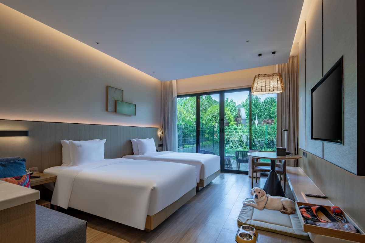 A hotel room with two beds, a desk, a TV, and a balcony with a view of greenery.