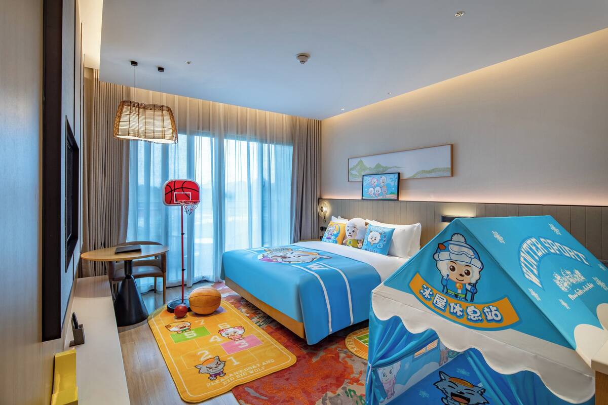 A hotel room with a large bed, a desk, a chair, a basketball, a mat with cartoon characters, and a ceiling fan.