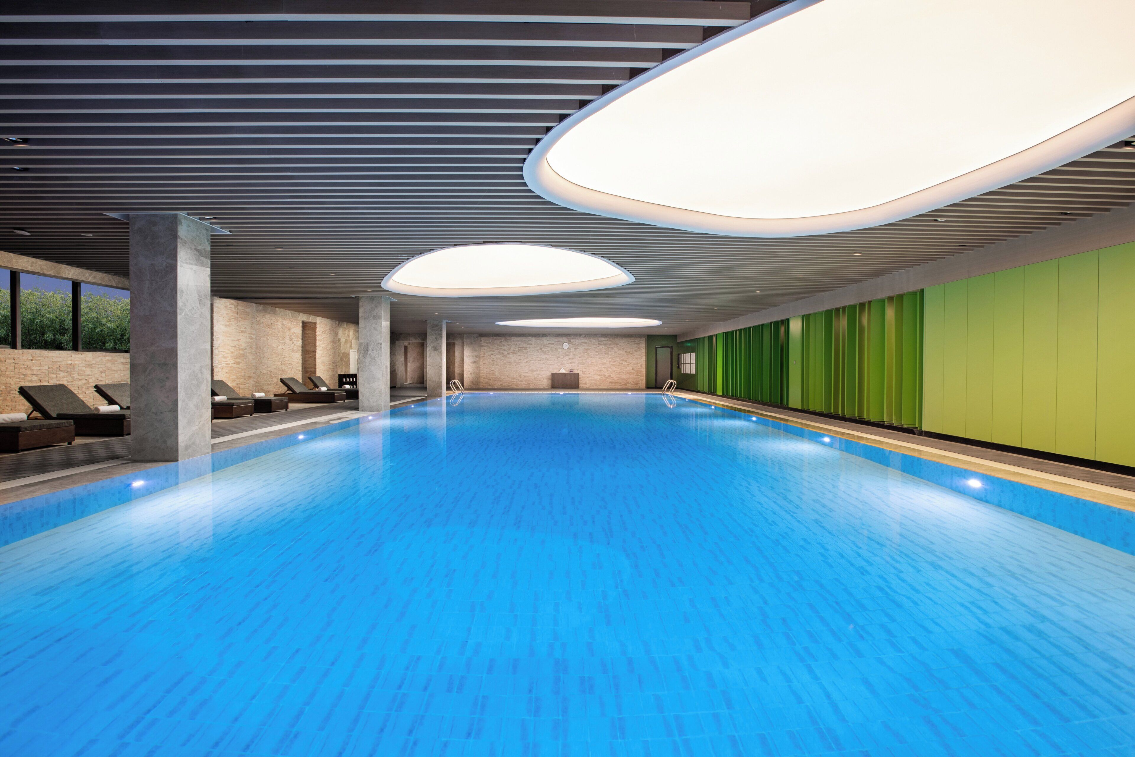 A large indoor swimming pool with a blue floor, surrounded by lounge chairs and a green wall.