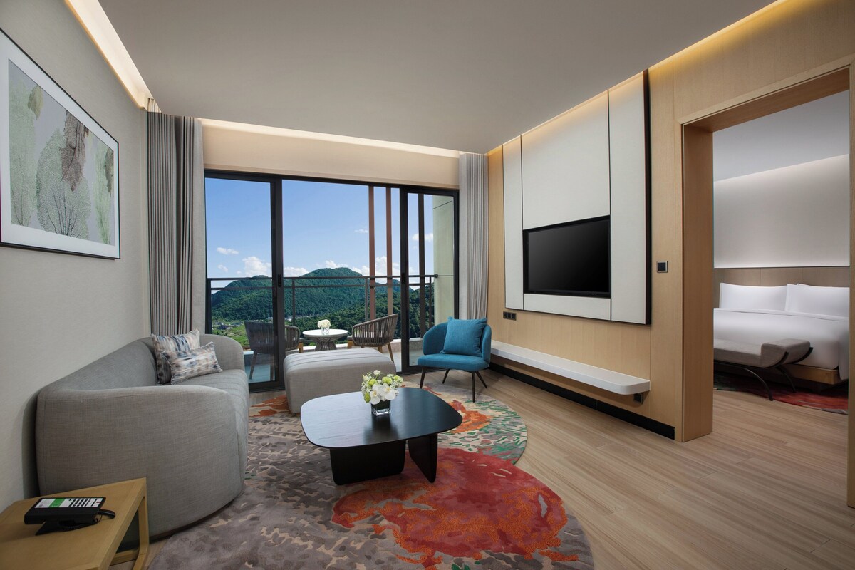 A modern hotel room with a large window, a flat-screen TV, a sofa, a coffee table, and a bed.